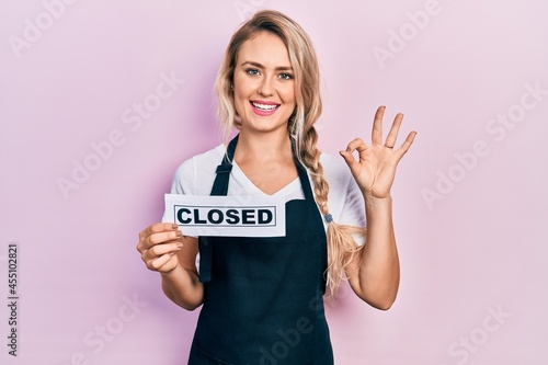 Beautiful young blonde woman wearing waitress apron holding closed banner doing ok sign with fingers  smiling friendly gesturing excellent symbol