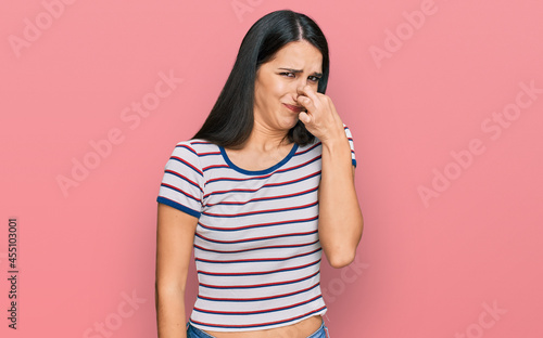 Young hispanic girl wearing casual striped t shirt smelling something stinky and disgusting, intolerable smell, holding breath with fingers on nose. bad smell