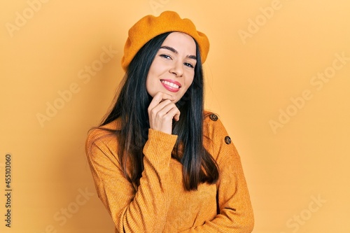 Young brunette woman wearing french look with beret smiling looking confident at the camera with crossed arms and hand on chin. thinking positive.