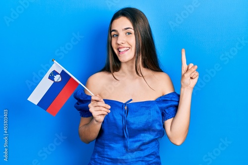 Young brunette teenager holding slovenia flag smiling with an idea or question pointing finger with happy face  number one