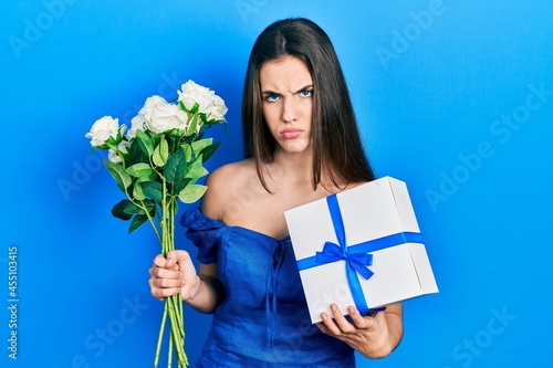 Young brunette teenager holding anniversary present and bouquet of flowers skeptic and nervous, frowning upset because of problem. negative person.