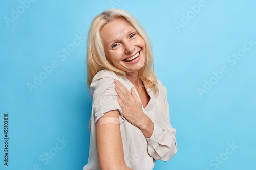 Fototapete Indoor shot of blonde senior woman gets vaccinated against coronavirus wears adhesive bandage on shoulder recieves vaccine shot isolated over blue background