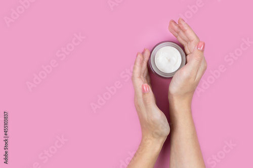Fototapeta Naklejka Na Ścianę i Meble -  A jar of white cream in the hands of a woman on a pink background. Place for a logo. Face cream or skin care products.