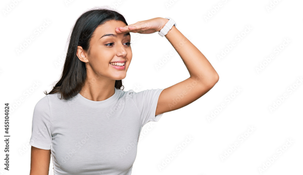 Young hispanic girl wearing casual white t shirt very happy and smiling looking far away with hand over head. searching concept. Young hispanic girl wearing casual white t shirt very happy and smiling looking far away with hand over head. searching concept.