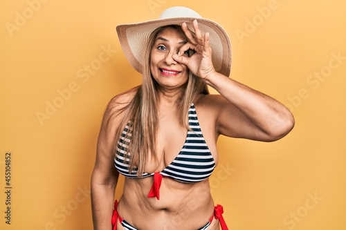 Middle age hispanic woman wearing bikini and summer hat doing ok gesture with hand smiling  eye looking through fingers with happy face.