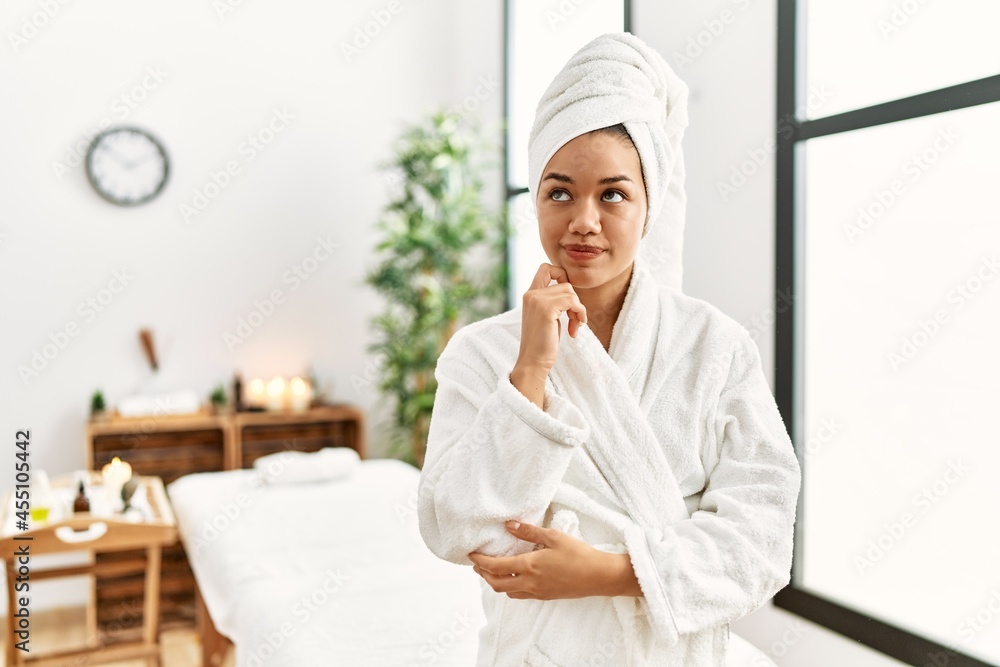 Young brunette woman wearing towel and bathrobe standing at beauty center with hand on chin thinking about question, pensive expression. smiling with thoughtful face. doubt concept. Young brunette woman wearing towel and bathrobe standing at beauty center with hand on chin thinking about question, pensive expression. smiling with thoughtful face. doubt concept.