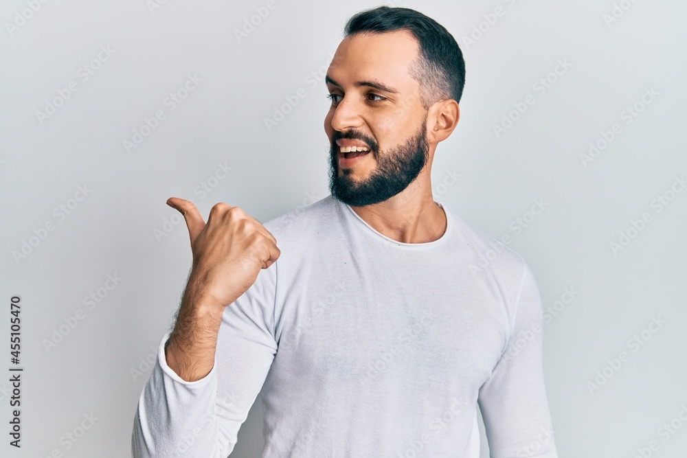 Young man with beard wearing casual white shirt pointing thumb up to the side smiling happy with open mouth Young man with beard wearing casual white shirt pointing thumb up to the side smiling happy with open mouth