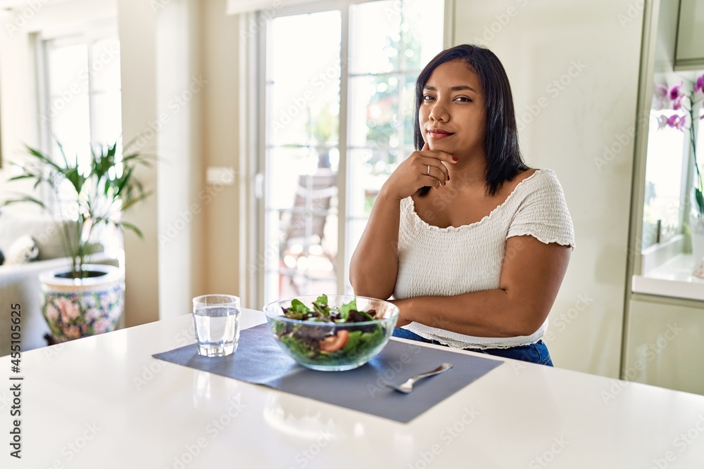Young hispanic woman eating healthy salad at home looking confident at the camera with smile with crossed arms and hand raised on chin. thinking positive.