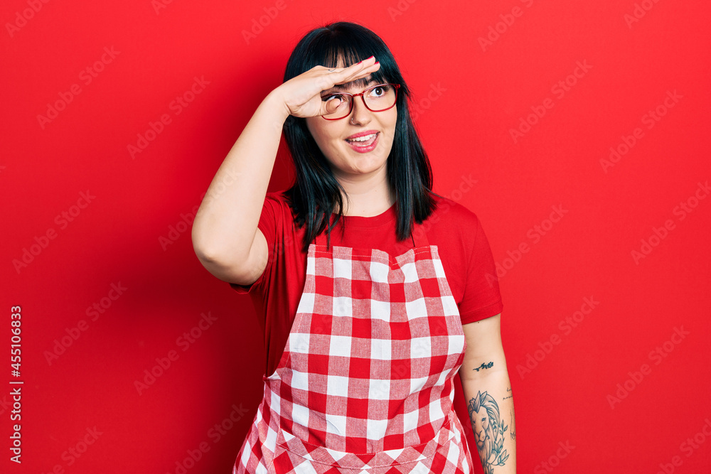 Young hispanic woman wearing cook apron and glasses very happy and smiling looking far away with hand over head. searching concept. Young hispanic woman wearing cook apron and glasses very happy and smiling looking far away with hand over head. searching concept.