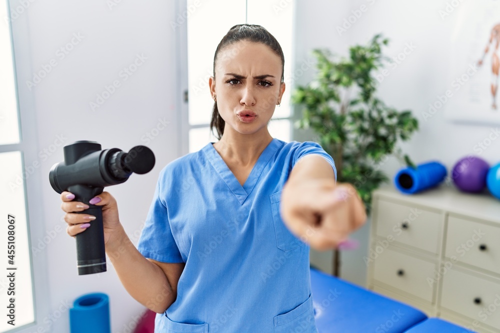 Young physiotherapist woman holding therapy massage gun at wellness center pointing with finger to the camera and to you, confident gesture looking serious Young physiotherapist woman holding therapy massage gun at wellness center pointing with finger to the camera and to you, confident gesture looking serious