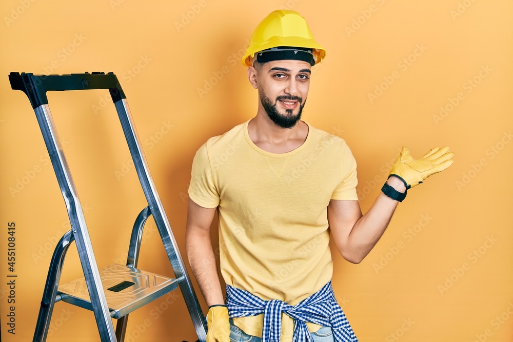 Handsome man with beard by construction stairs wearing hardhat smiling cheerful presenting and pointing with palm of hand looking at the camera. Handsome man with beard by construction stairs wearing hardhat smiling cheerful presenting and pointing with palm of hand looking at the camera.