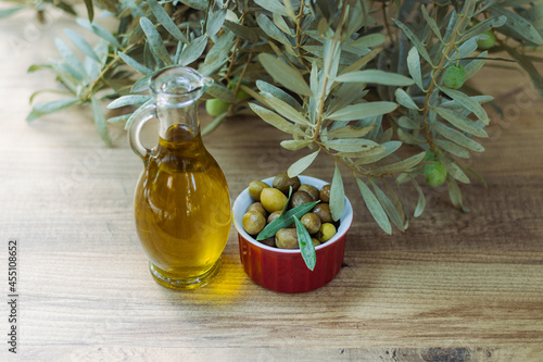 Closeup photo of green olives in a red bowl next to an olive oil bottle and together with an olive tree leaf
