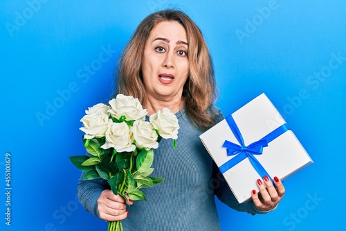 Middle age caucasian woman holding anniversary present and bouquet of flowers clueless and confused expression. doubt concept.