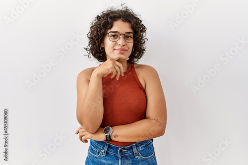 Young hispanic woman wearing glasses standing over isolated background looking confident at the camera smiling with crossed arms and hand raised on chin. thinking positive.