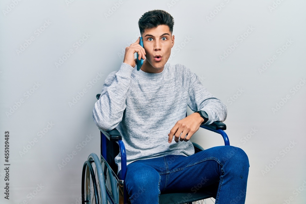 Young hispanic man sitting on wheelchair talking on the smartphone scared and amazed with open mouth for surprise, disbelief face Young hispanic man sitting on wheelchair talking on the smartphone scared and amazed with open mouth for surprise, disbelief face