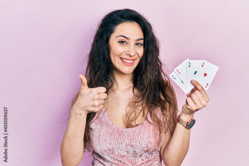 Young hispanic woman holding poker cards smiling happy and positive, thumb up doing excellent and approval sign Young hispanic woman holding poker cards smiling happy and positive, thumb up doing excellent and approval sign