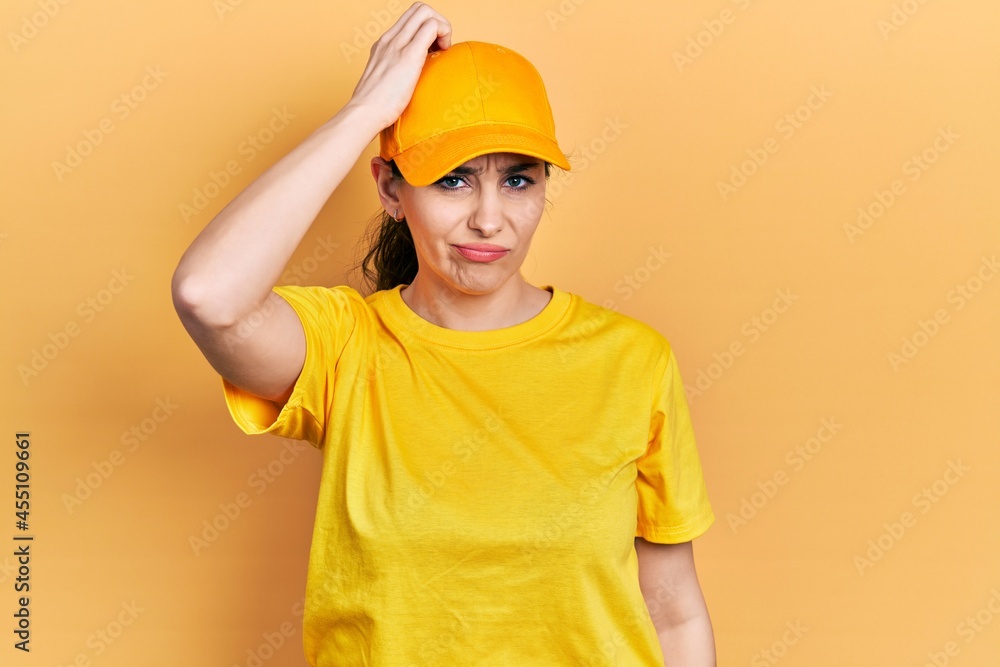 Young hispanic woman wearing delivery uniform and cap confuse and wonder about question. uncertain with doubt, thinking with hand on head. pensive concept.