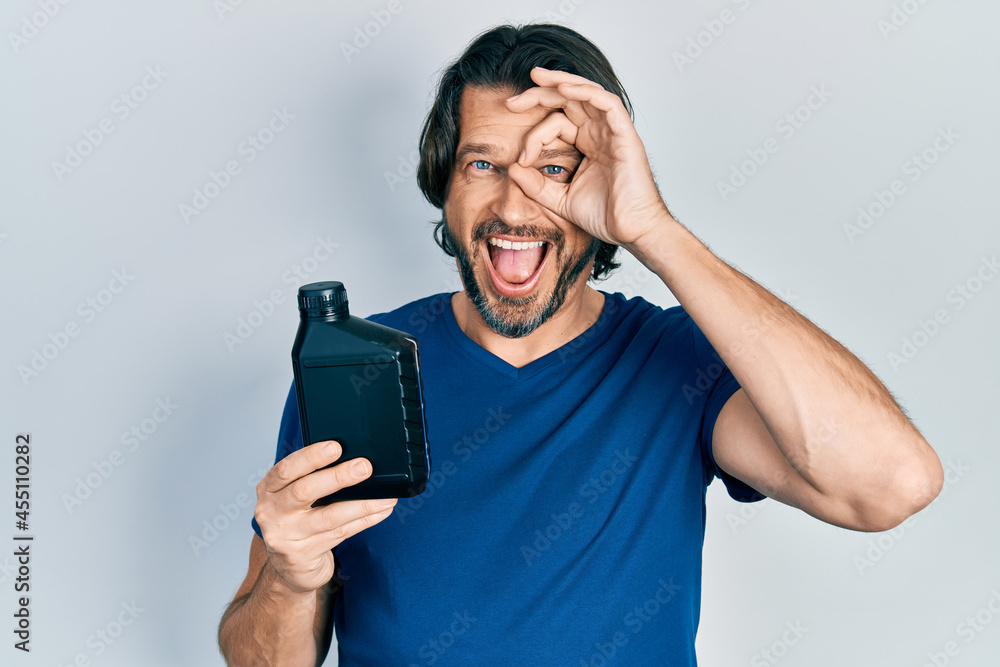 Middle age caucasian man holding motor oil bottle smiling happy doing ok sign with hand on eye looking through fingers Middle age caucasian man holding motor oil bottle smiling happy doing ok sign with hand on eye looking through fingers