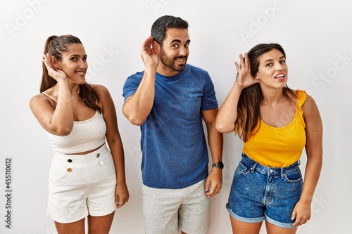 Group of young hispanic people standing over isolated background smiling with hand over ear listening an hearing to rumor or gossip. deafness concept.