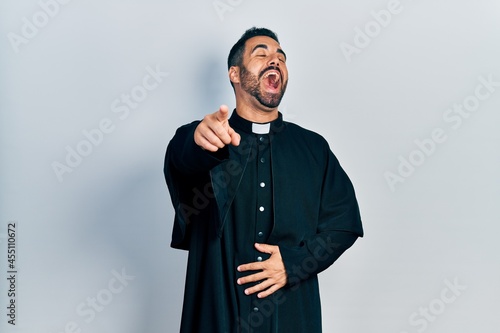 Handsome hispanic man with beard wearing catholic priest robe laughing at you, pointing finger to the camera with hand over body, shame expression
