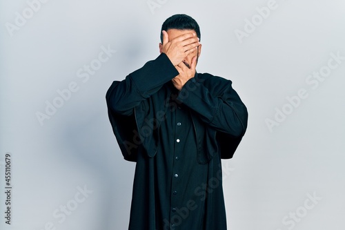 Handsome hispanic man with beard wearing catholic priest robe covering eyes and mouth with hands, surprised and shocked. hiding emotion