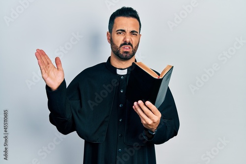 Handsome hispanic priest man with beard holding bible and christian cross clueless and confused expression. doubt concept.