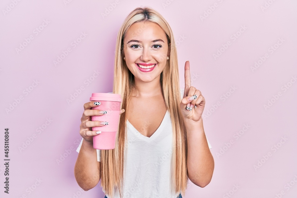 Young blonde girl holding coffee smiling with an idea or question pointing finger with happy face, number one Young blonde girl holding coffee smiling with an idea or question pointing finger with happy face, number one