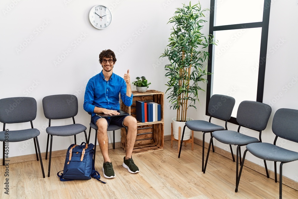 Fototapeta premium Young hispanic man sitting at waiting room with folder smiling happy and positive, thumb up doing excellent and approval sign