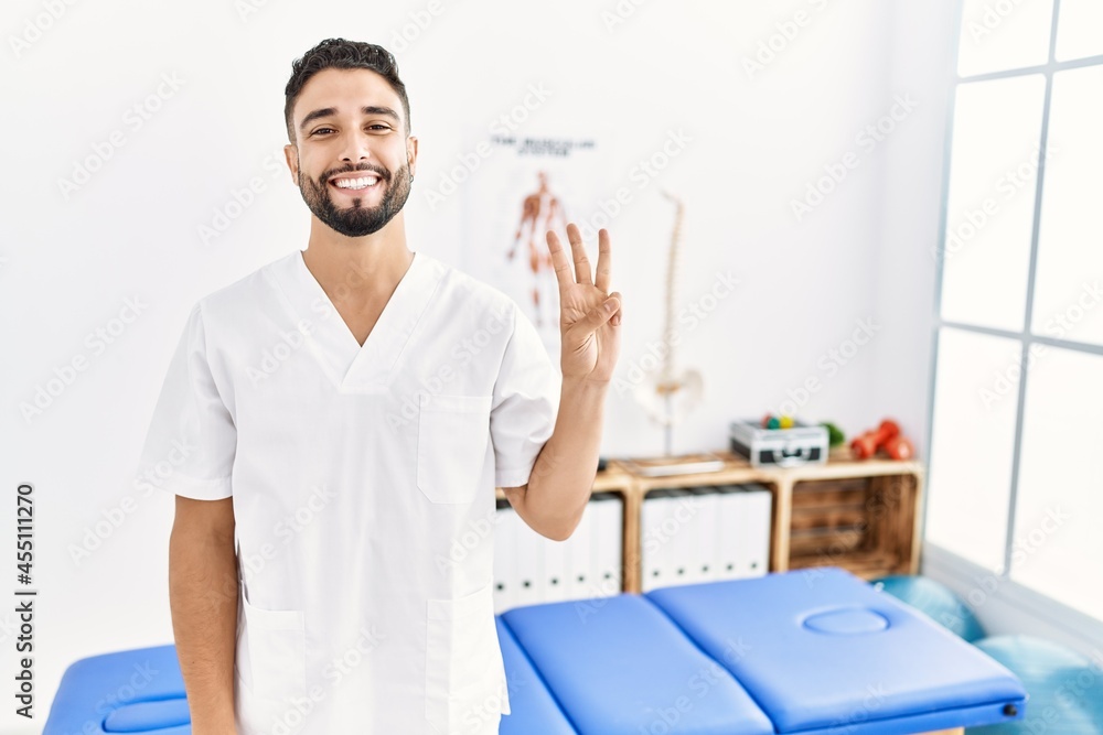 Young handsome man with beard working at pain recovery clinic showing and pointing up with fingers number three while smiling confident and happy. Young handsome man with beard working at pain recovery clinic showing and pointing up with fingers number three while smiling confident and happy.