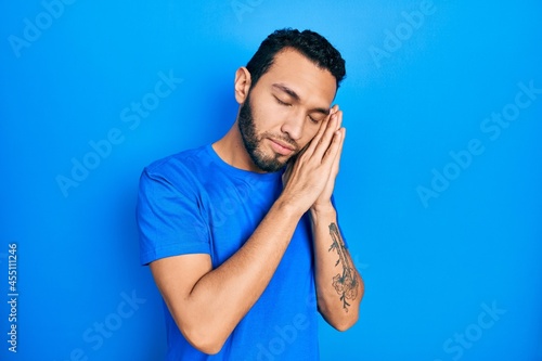 Hispanic man with beard wearing casual blue t shirt sleeping tired dreaming and posing with hands together while smiling with closed eyes.
