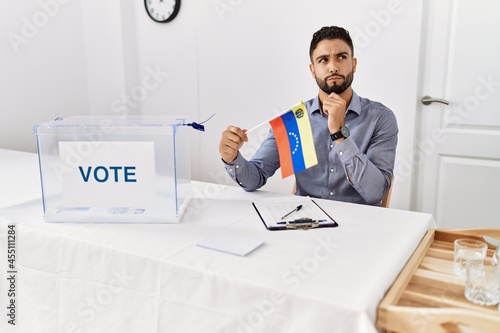 Young handsome man with beard at political campaign election holding venezuela flag serious face thinking about question with hand on chin, thoughtful about confusing idea