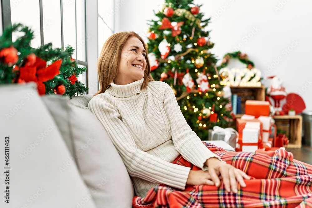 Middle age woman sitting on the sofa by christmas tree looking away to side with smile on face, natural expression. laughing confident. Middle age woman sitting on the sofa by christmas tree looking away to side with smile on face, natural expression. laughing confident.