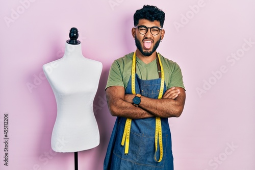 Arab man with beard dressmaker designer wearing atelier apron sticking tongue out happy with funny expression.