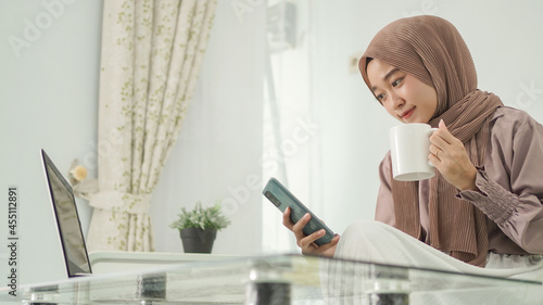 beautiful woman in hijab working from home looking for good ideas on her smartphone while enjoying a drink