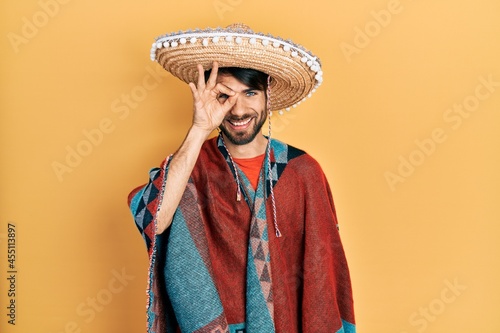 Young hispanic man holding mexican hat doing ok gesture with hand smiling, eye looking through fingers with happy face.