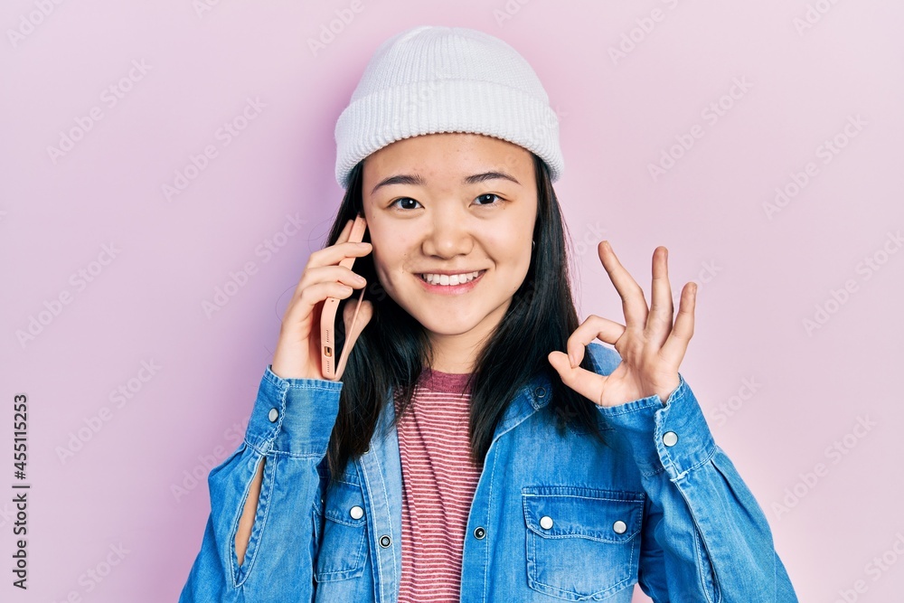 Young chinese girl having conversation talking on the smartphone doing ok sign with fingers, smiling friendly gesturing excellent symbol Young chinese girl having conversation talking on the smartphone doing ok sign with fingers, smiling friendly gesturing excellent symbol