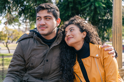 Young couple sitting in a park while the man hugs the woman, she looks at him. © HC FOTOSTUDIO