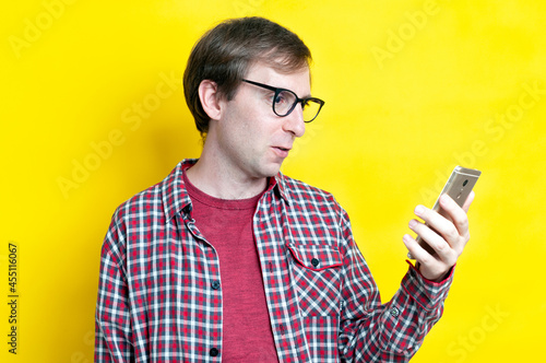 handsome surprised man in red checkered shirt and glasses looking at smartphone on yellow background with copy space