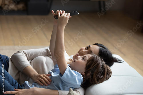 Happy little kid boy making selfie photo on web camera with caring affectionate young korean multiracial mother, recording video, streaming stories in social network, lying together on cozy sofa.