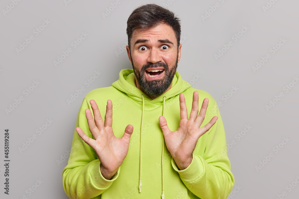 Fototapeta premium Scared panicking bearded man looks frightened has phobias raises palm tries to protect himself from something awful wears casual hoodie looks desperate and devastated isolated over grey background