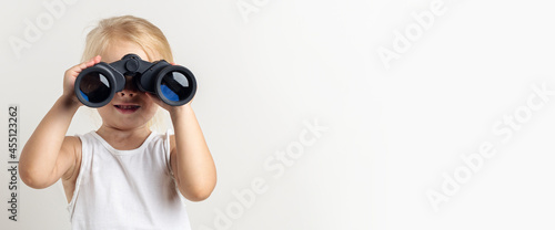 Smiling blonde child looks through binoculars on a light background in the studio. Banner © Alex