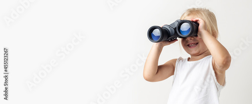 Smiling blonde child looks through binoculars on a light background in the studio. Banner