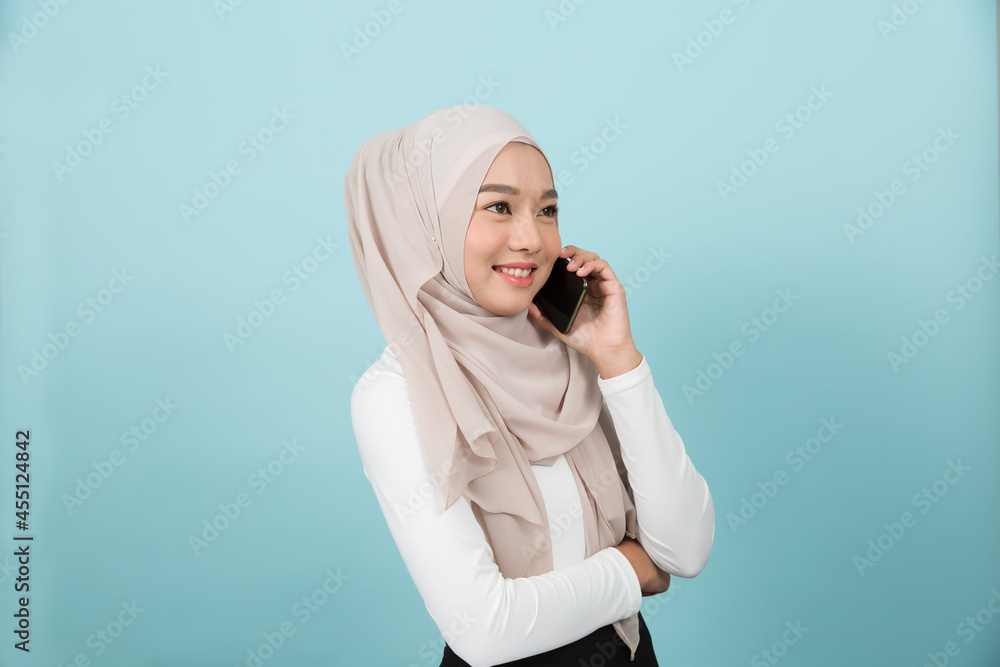 Portrait of smiling Asian young Muslim woman in hijab head scarf standing and using smart phone against blue colour background Portrait of smiling Asian young Muslim woman in hijab head scarf standing and using smart phone against blue colour background