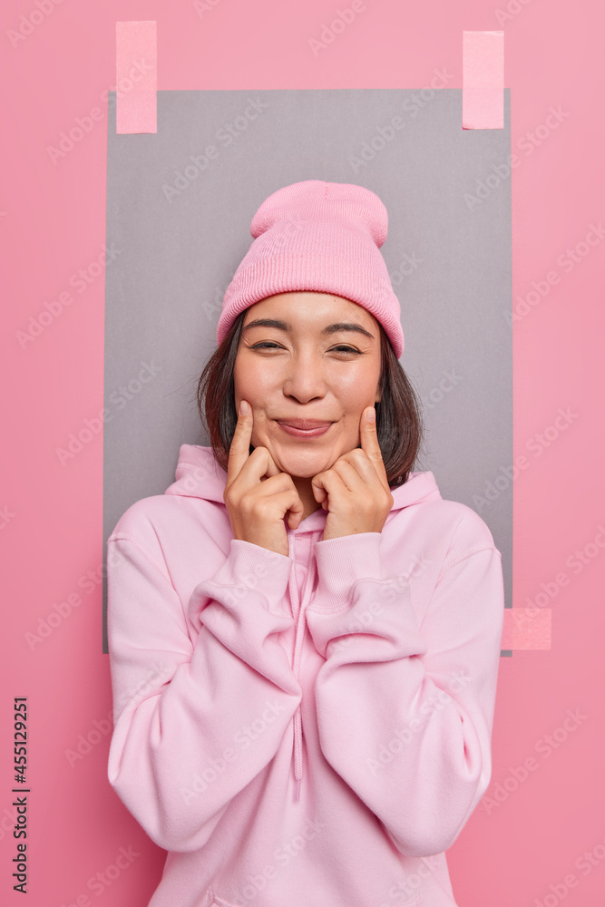 Pleased teenage girl forces smile keeps fingers near corner of lips has sincere positive expression dressed in casual hoodie poses indoor against blank copy space. Human emotions and feelings Pleased teenage girl forces smile keeps fingers near corner of lips has sincere positive expression dressed in casual hoodie poses indoor against blank copy space. Human emotions and feelings