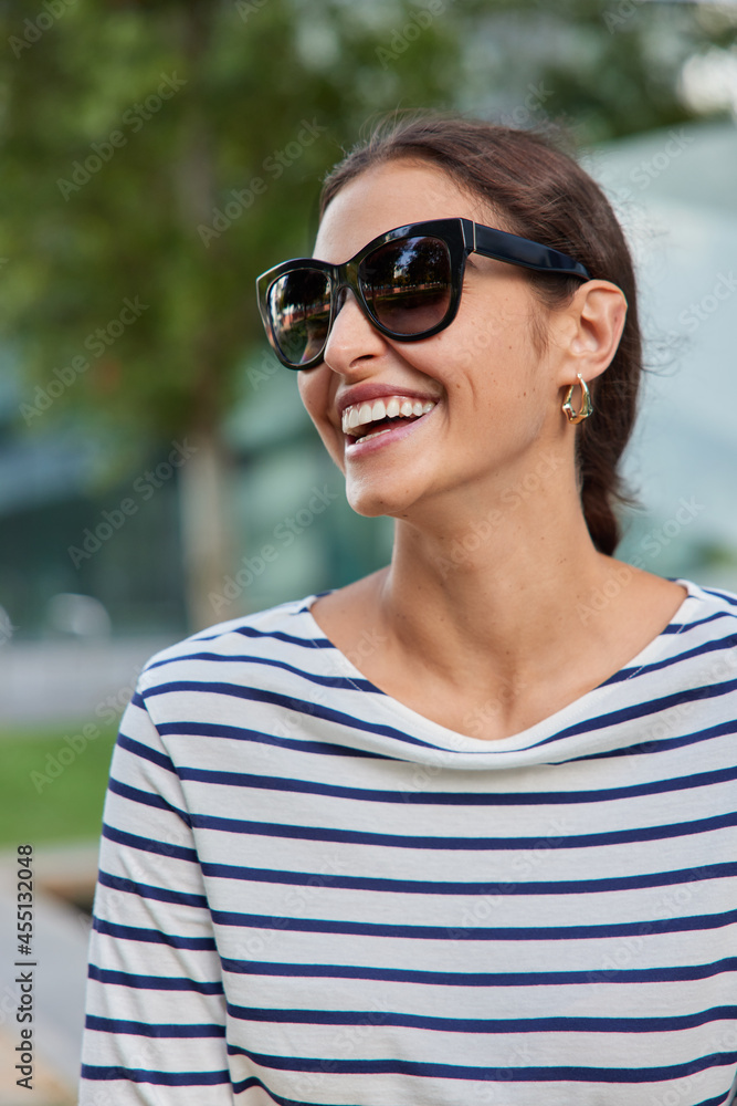 Vertical outdoor shot of happy adult woman poses at street relaxes in park wears sunglasses striped jumper enjoys spring day meets with friend. People lifestyle leisure and emotions concept. Vertical outdoor shot of happy adult woman poses at street relaxes in park wears sunglasses striped jumper enjoys spring day meets with friend. People lifestyle leisure and emotions concept.