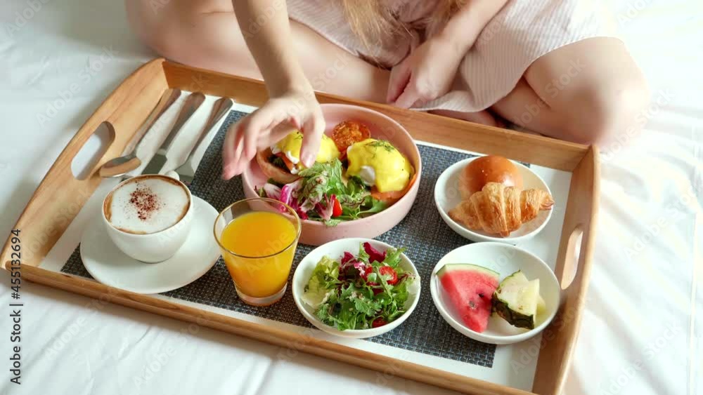 Breakfast in Bed - Female Hand Put a Glass of Orange Juice on Wooden Tray with Fresh Food, Eggs, Fruits, Cup of Coffee. Room Service in Hotel on Vacation. Woman Sitting on Bed and Eat Her Morning Food Breakfast in Bed - Female Hand Put a Glass of Orange Juice on Wooden Tray with Fresh Food, Eggs, Fruits, Cup of Coffee. Room Service in Hotel on Vacation. Woman Sitting on Bed and Eat Her Morning Food