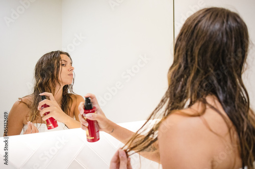 young beautiful millennial brunette white woman at home no makeup wet hair after shower looking at herself in the bathroom mirror applying heat protection spray on her hair self care routine beauty photo