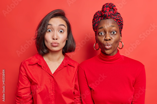 Diverse women best friends stand closely to each other keep lips rounded wait for kiss wears red clothes pose against bright studio wall. Mixed race female models with puckered lips. Face expressions