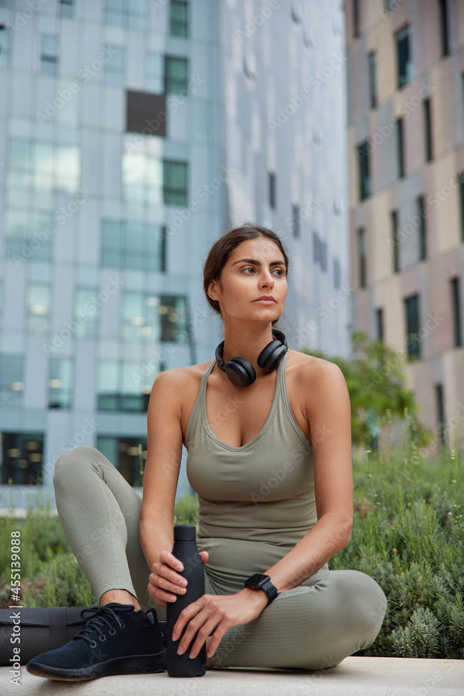 Fototapeta premium Pensive young woman gets ready for running routine holds bottle of fresh water feels thirsty after cardio training dressed in activewear poses against cityscrapers outdoor. Sport activity in open air