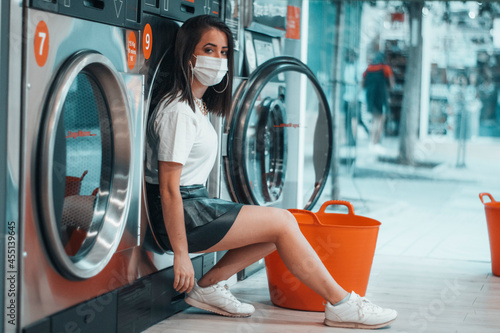 Muchacha joven de pelo castaño con mascarilla posando en la tienda de la lavanderia junto a la cesta de ropa para limpiar las prendas en la lavadora del comercio